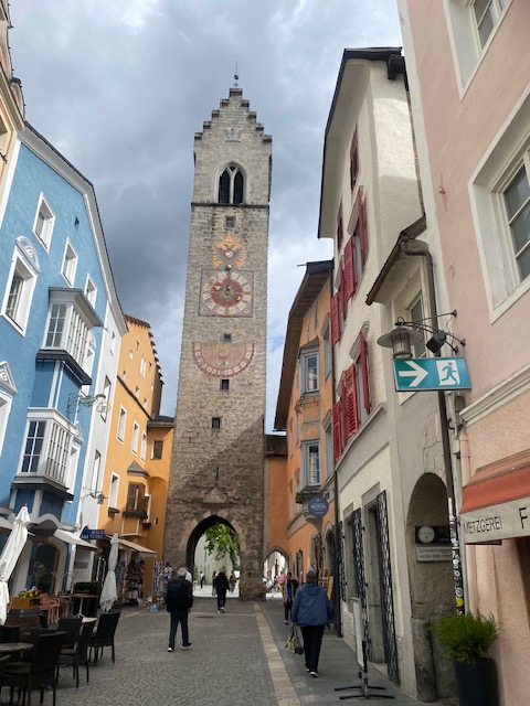 A view along a colourful cobbled street toward the medieval Zwölferturm clock tower in Sterzing (Vipiteno), South Tyrol, framed by pastel façades beneath a dramatic, cloud-filled sky.